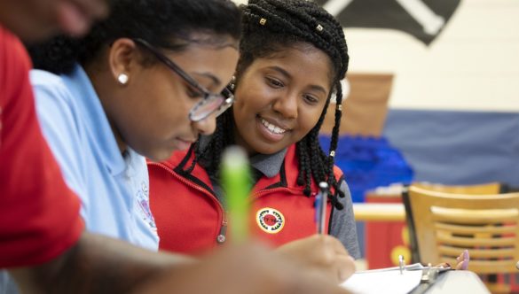 two students are doing independent work at a table while an AmeriCorps member looks at their work and smiles