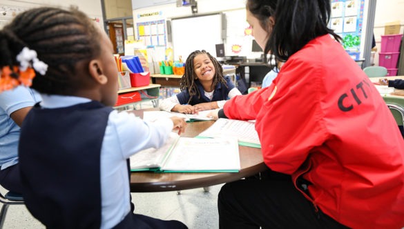 two young students, one smiling in a classroom as a City Year corps member teaches them around a table in a classroom