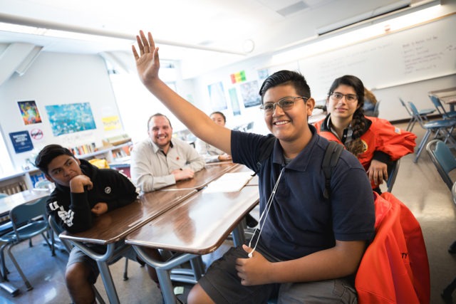 A student raising his hand as he sits with other students and city year americorps members in a classroom