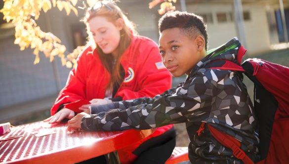City year americorps member and a student with a backpack sitting at a table in a school courtyard in the sunshine