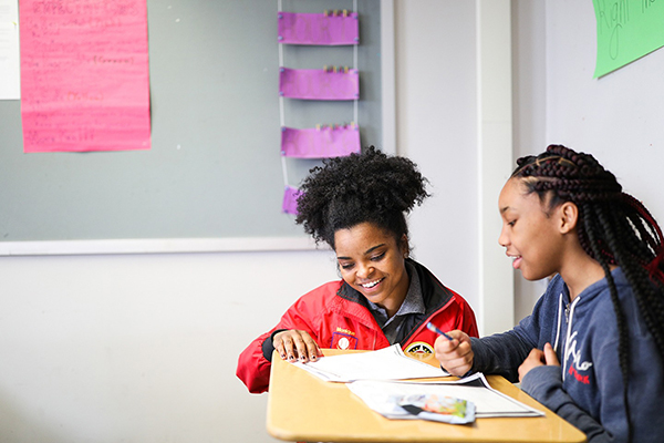 City Year AmeriCorps member kneeling next to a student at a desk as they review work