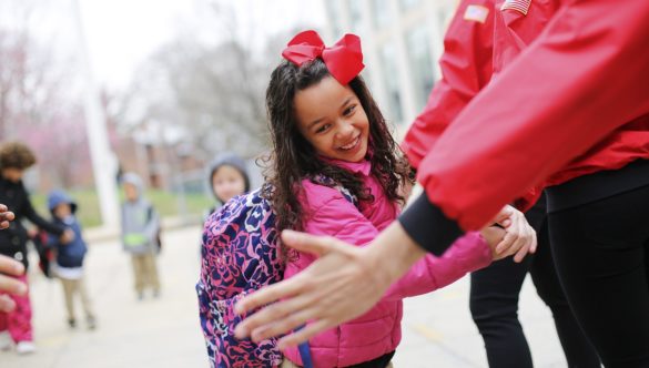 An elementary school student wearing a bow and a backpack high-fives several AmeriCorps members as she arrives at school on time
