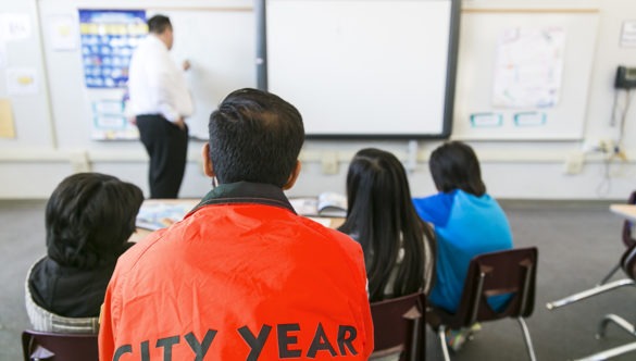 A city year americorps member looks on from the back of a classroom as a teacher writes on a board and students look on