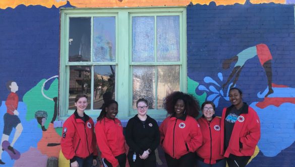 AmeriCorps members stand in front of a colorful mural with their red jackets on.