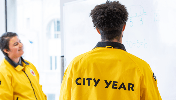 two City Year AmeriCorps members in yellow jackets face a whiteboard with math problems on it